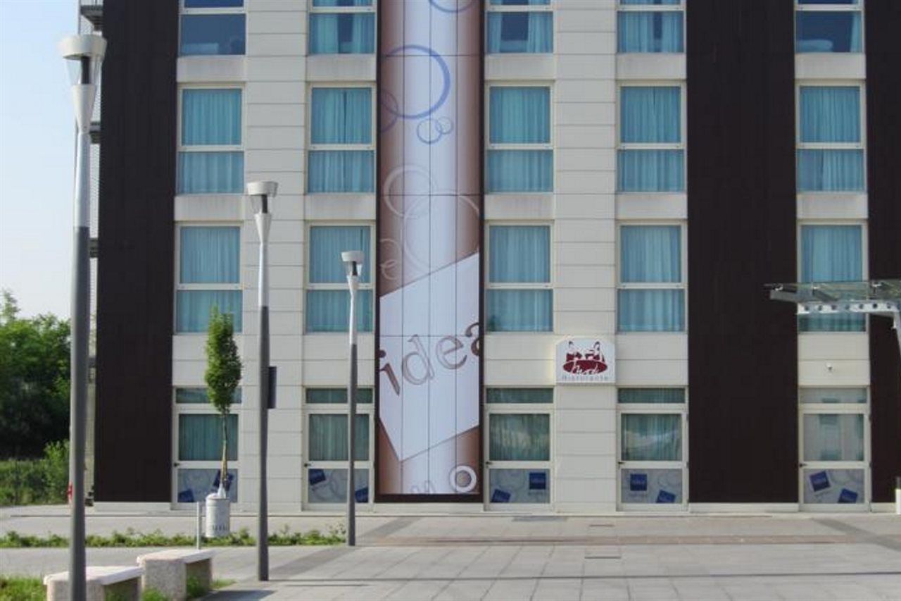 Idea Hotel Milano Malpensa Airport Case Nuove  Eksteriør billede The photo shows a modern building facade. The exterior has a mix of white and dark panels, with several large windows featuring blue curtains. A prominent banner or advertisement runs vertically down the center of the building, showcasing an abstract