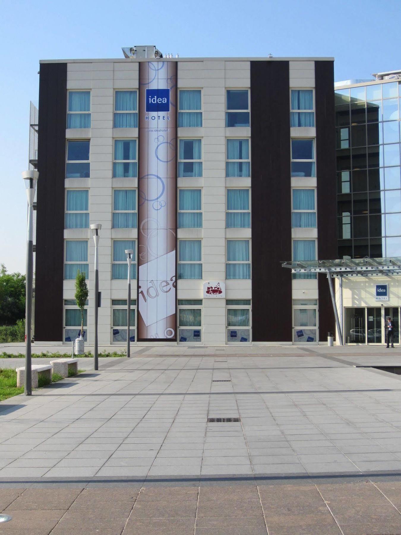 Idea Hotel Milano Malpensa Airport Case Nuove  Eksteriør billede The photo shows the facade of a multi-story building, likely a hotel. It features a combination of light and dark exterior panels. In the center, there is a large banner with a design, possibly advertising or displaying a logo. The entrance appears t