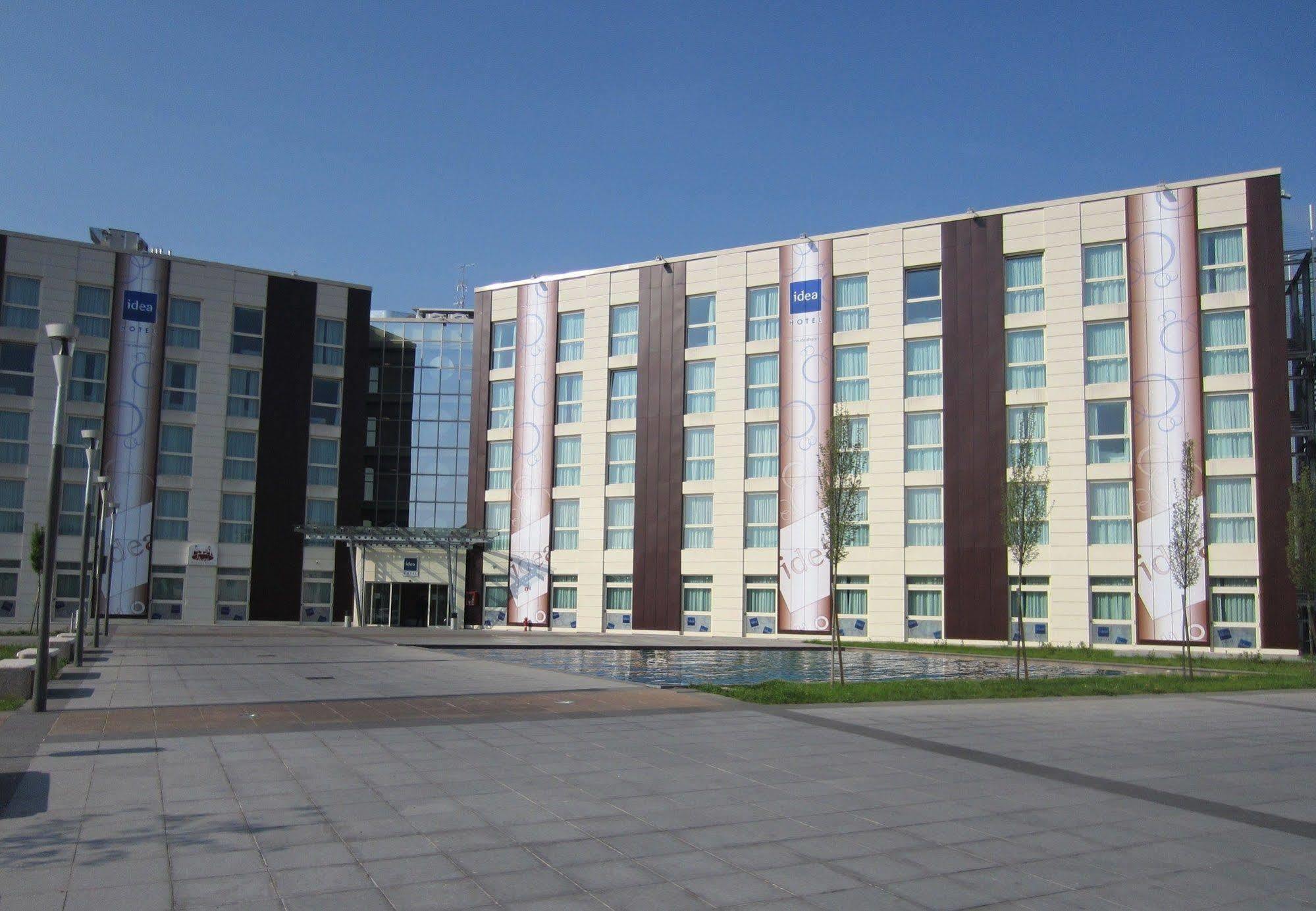 Idea Hotel Milano Malpensa Airport Case Nuove  Eksteriør billede The photo shows a modern multi-story building, likely a hotel or office complex. The building features a combination of light-colored and dark-colored panels, with large windows allowing for natural light. There are promotional banners displayed on t
