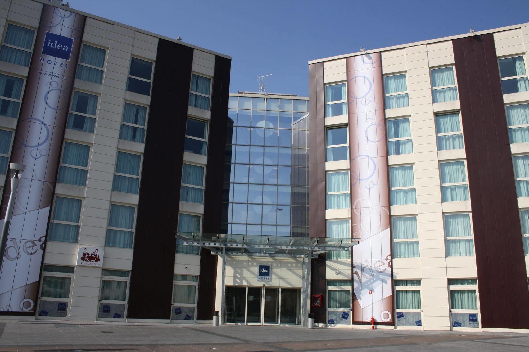 Idea Hotel Milano Malpensa Airport Case Nuove  Eksteriør billede The photo shows the exterior of a modern hotel building. It features a large glass entrance surrounded by two taller sections composed of light-colored and dark-colored panels. The left side has a banner displaying the hotel name, and there are large